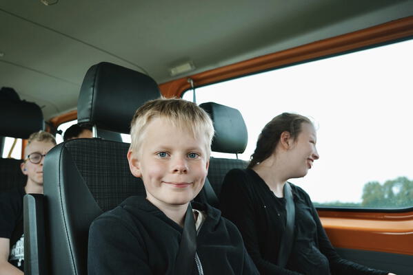 Ein lachender Junge sitzt in einem orangen Bus. Neben ihm eine Mitfahrerin, die lächelnd aus dem Fenster guckt. Hinter ihnen sitzen noch zwei Kinder
